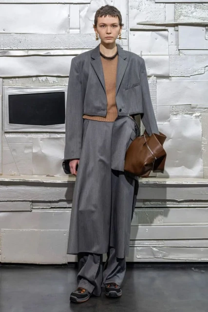 A model poses in a grey, oversized suit with a brown sweater and brown purse against a silver metal panel wall in a full-length studio shot.