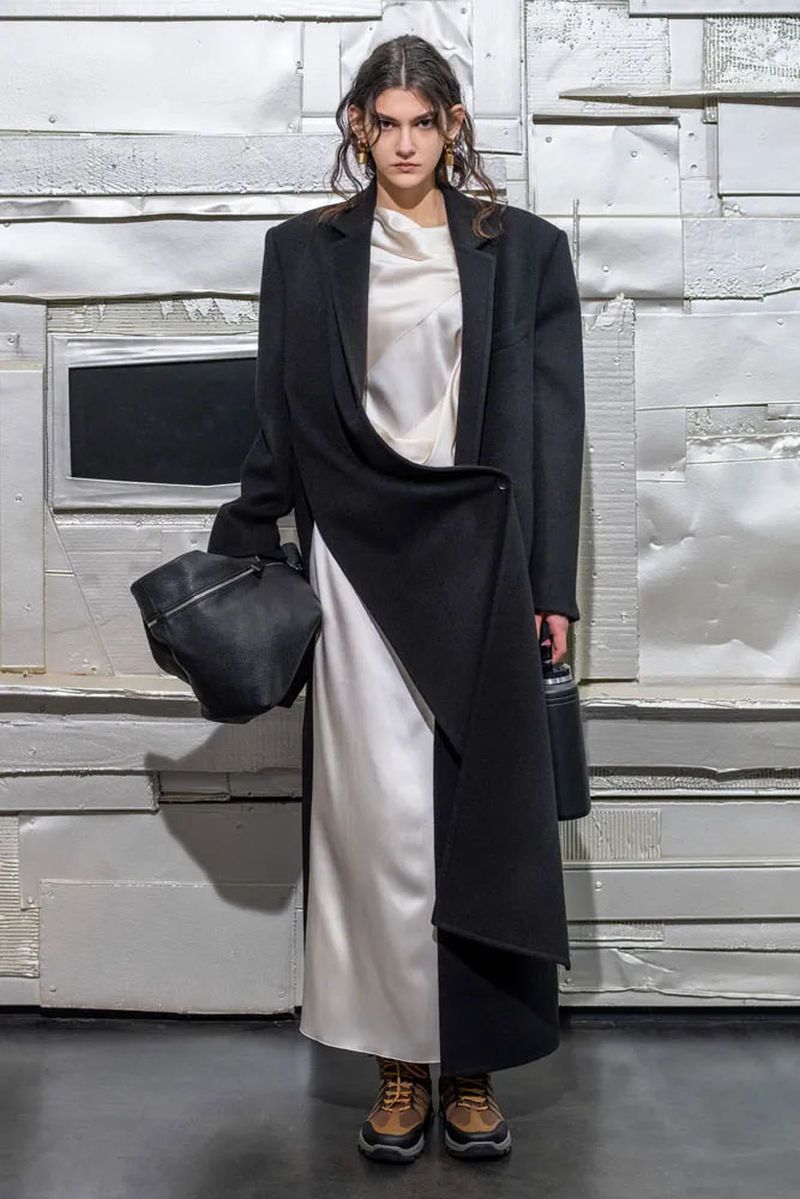 A model in a black coat and white dress poses against a distressed wall, holding a soft leather bag, in a studio fashion shot.