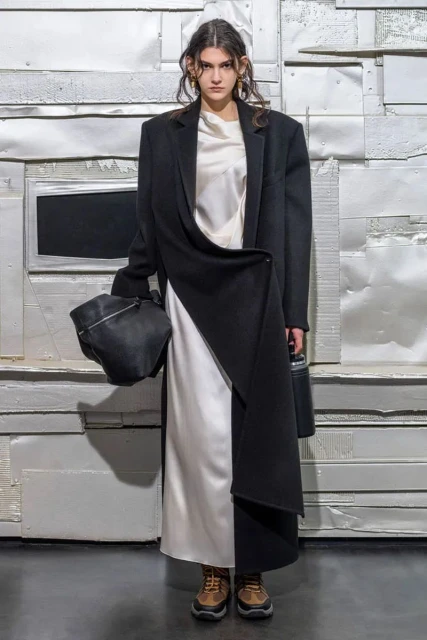 A model in a black coat and white dress poses against a distressed wall, holding a soft leather bag, in a studio fashion shot.