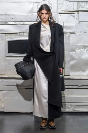 A model in a black coat and white dress poses against a distressed wall, holding a soft leather bag, in a studio fashion shot.