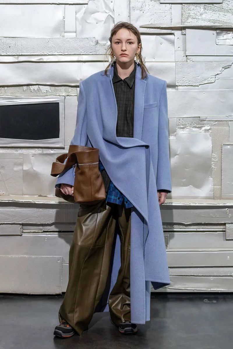 A woman in a long blue coat, wide brown pants, and sneakers stands in front of a textured white background, holding a brown leather handbag.