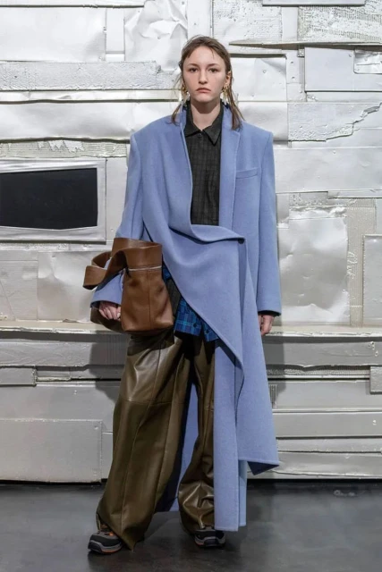 A woman in a long blue coat, wide brown pants, and sneakers stands in front of a textured white background, holding a brown leather handbag.