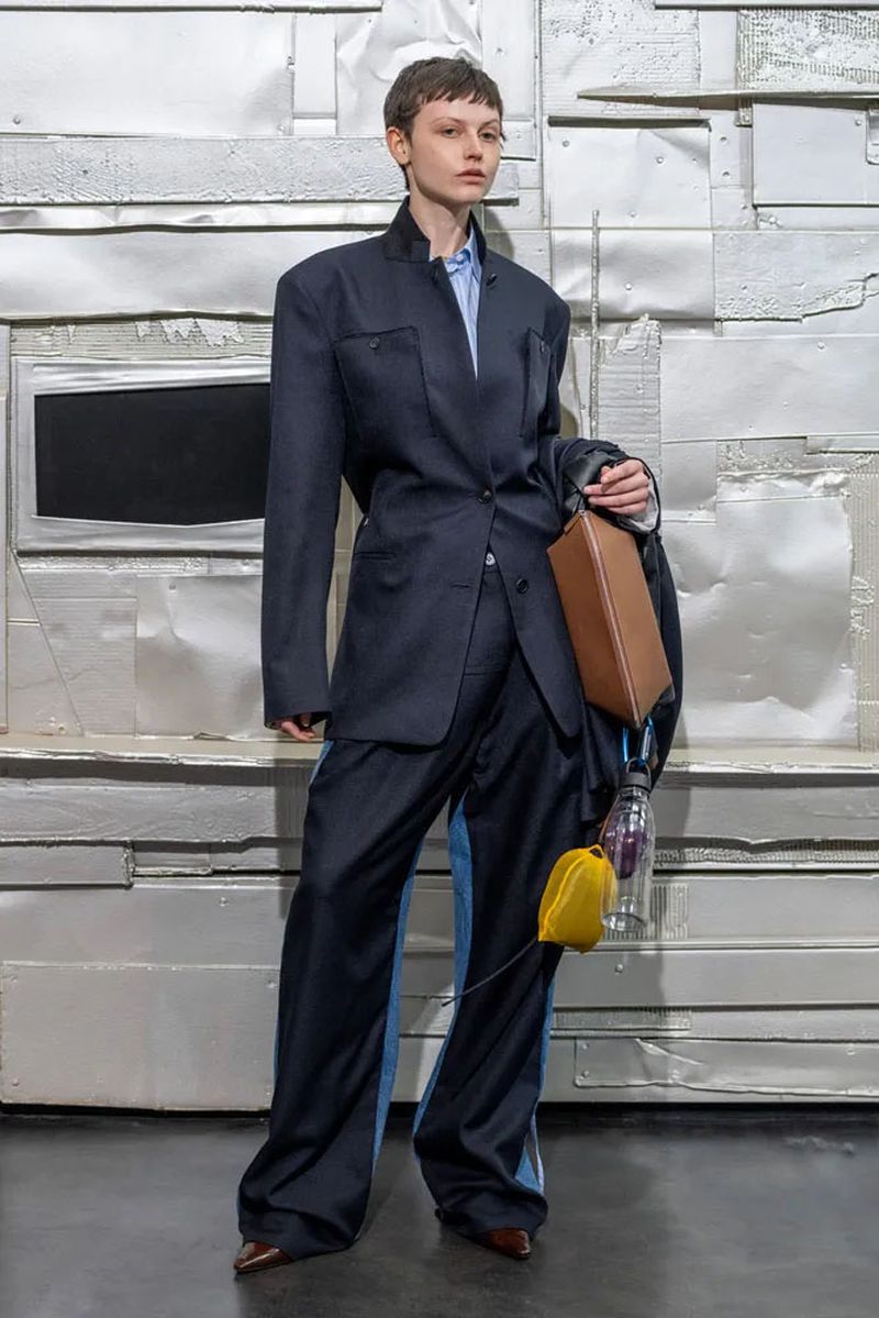 A model poses in a dark blue oversized suit with wide-legged pants, a light blue shirt, a brown purse, and a yellow coin purse against a silver background.