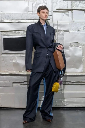 A model poses in a dark blue oversized suit with wide-legged pants, a light blue shirt, a brown purse, and a yellow coin purse against a silver background.