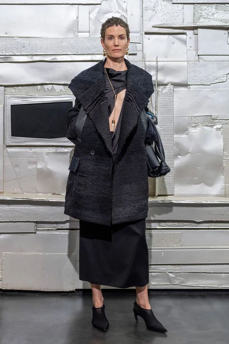 A full shot of a woman in a black coat and dress stands against a silver wall. The lighting is bright and even, capturing her neutral expression.