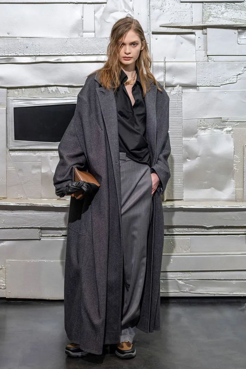 A woman with long hair poses in a long, dark gray coat, gray pants, black top, and brown bag against a white textured background.