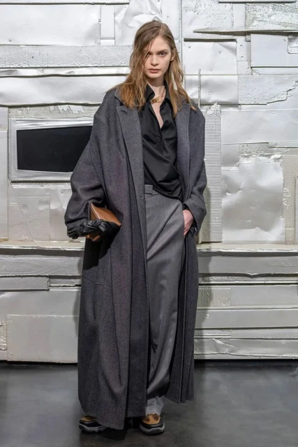 A woman with long hair poses in a long, dark gray coat, gray pants, black top, and brown bag against a white textured background.