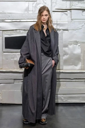 A woman with long hair poses in a long, dark gray coat, gray pants, black top, and brown bag against a white textured background.