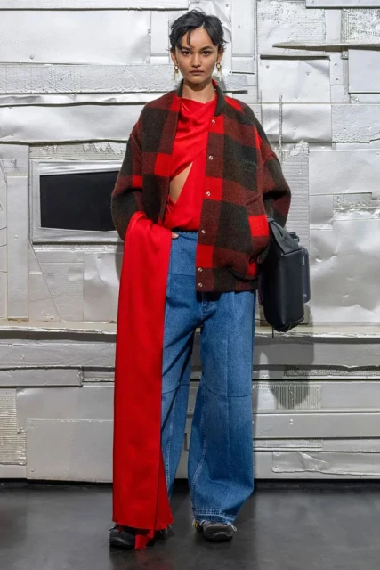 Full-length shot of a young woman in wide-legged jeans, a red top, and a buffalo plaid jacket, with a black bag and a silver background.