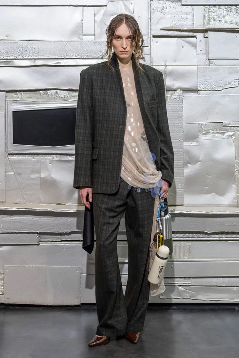 Full-length shot of androgynous adult in a plaid suit, sheer blouse, and brown heels standing against a silver metallic textured background.
