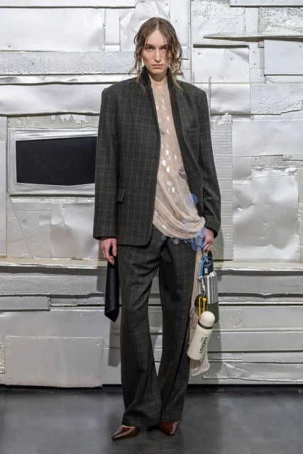 Full-length shot of androgynous adult in a plaid suit, sheer blouse, and brown heels standing against a silver metallic textured background.