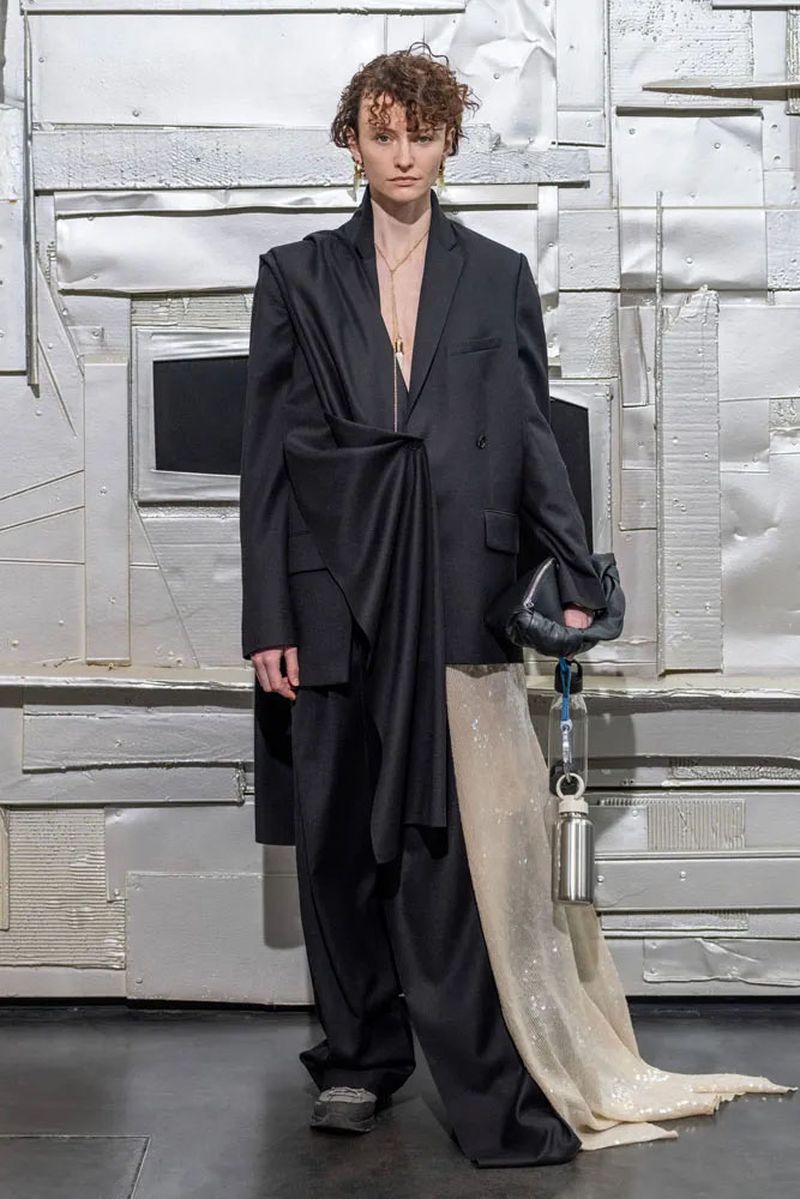 Woman poses in an oversized black blazer and matching pants with a textured silver backdrop. She holds a black clutch.