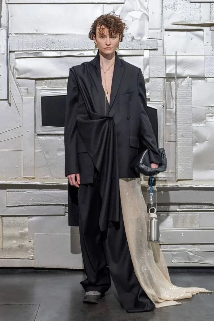 Woman poses in an oversized black blazer and matching pants with a textured silver backdrop. She holds a black clutch.