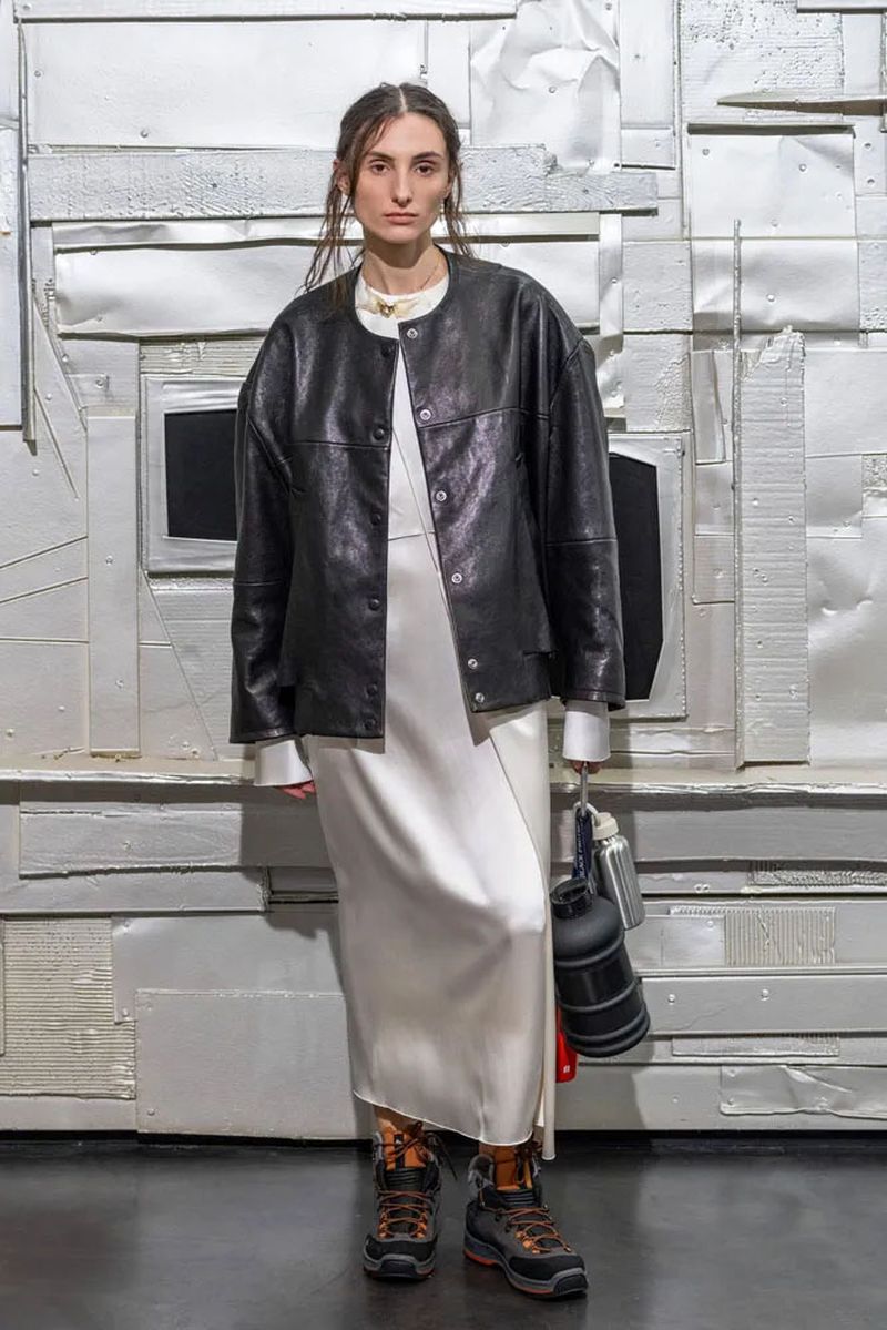 Adult female wearing a white dress and black jacket stands in a studio with a textured silver metallic background and black flooring.