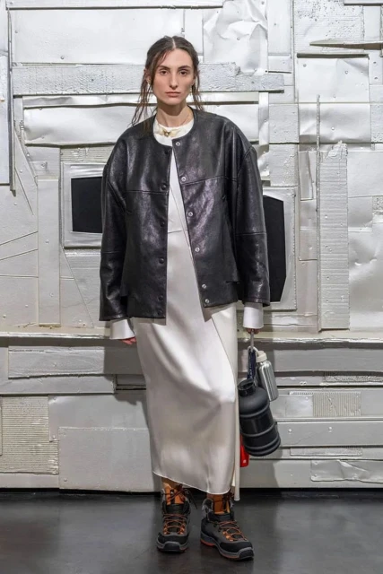 Adult female wearing a white dress and black jacket stands in a studio with a textured silver metallic background and black flooring.