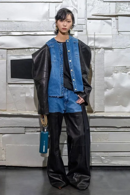 Full-length shot of a woman in a denim vest and leather pants, accessorized with a water bottle, posing in front of a silver background.
