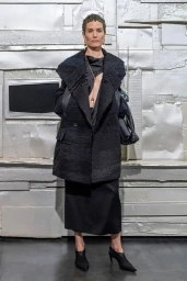 Model standing in a structured black coat and matching skirt against a textured metallic wall background.