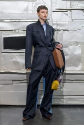 Model wearing an oversized navy tailored suit with wide-leg trousers, carrying a tan leather bag against a textured metallic backdrop.