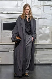 Model wearing an oversized charcoal maxi coat, black collared shirt, and grey trousers holding a brown clutch against a textured wall.