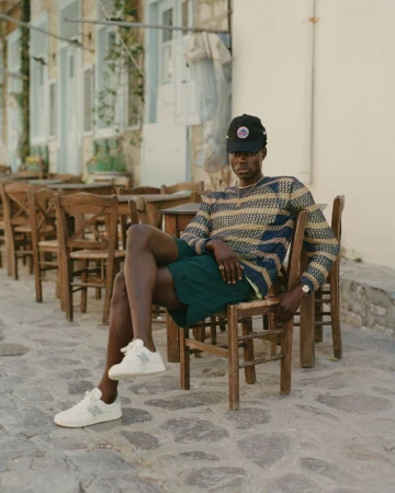 Man in sweater, shorts, sneakers, and baseball cap sitting on a wooden chair outside a building with shuttered windows on a stone surface.