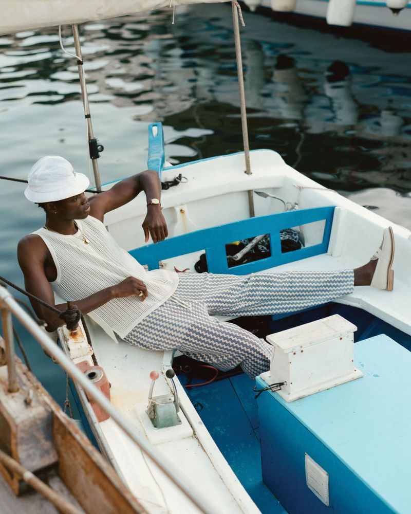 Young adult in summer fashion, lounging in a small boat on the water with white hat, knit top, patterned pants and a wristwatch.