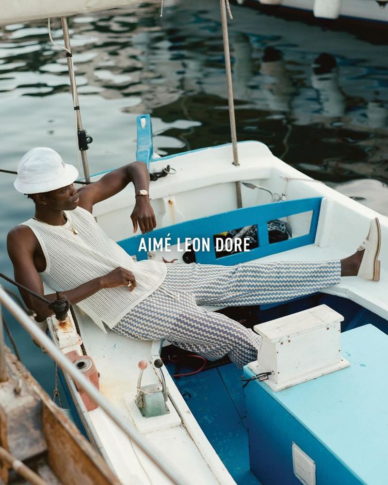 Stylish adult male model in sleeveless top, patterned trousers, and hat relaxes on a boat in a daytime outdoor fashion editorial.