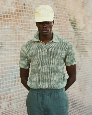 Young adult Black male wearing a yellow baseball cap, patterned polo shirt, and shorts stands against a textured neutral wall.