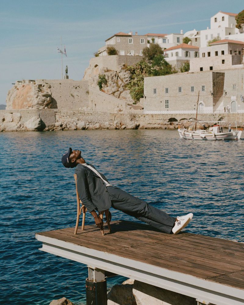 A dark-skinned adult man in a suit and baseball cap is reclining on a chair on a wooden dock, with an ocean and a coastal town in the background.