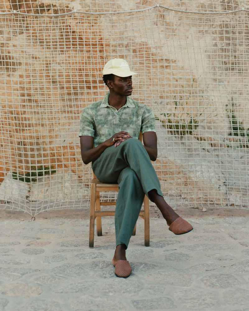 A fashionable Black man sits on a chair outside, wearing a cap, green patterned shirt, green trousers, and loafers, posing thoughtfully.