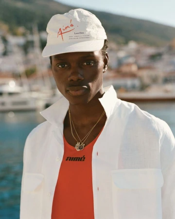 Close-up shot of an adult Black male model wearing a white cap, gold necklaces, and a red shirt with a white button-down overshirt outdoors.