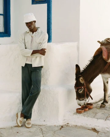 A man in a jacket and pants stands next to a donkey with a saddle eating from bowls on white steps near a building with a blue window.
