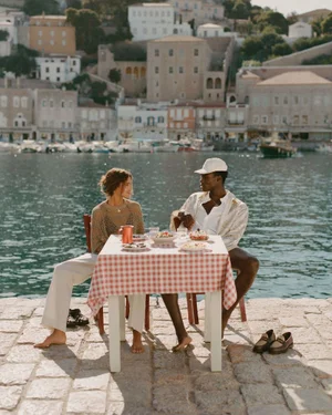 Man and woman sitting at a pink gingham dining table on a stone quay by the harbor during the Aimé Leon Dore Hydra SS24 shoot.