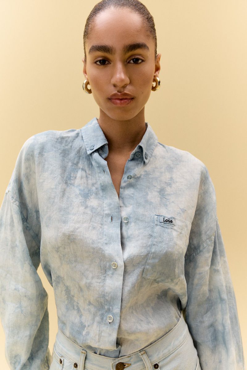 Close-up of a woman in a light blue tie-dye button-down shirt and light wash jeans against a light yellow background. She wears gold hoops.