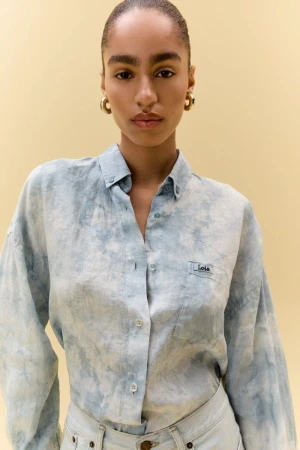 Model Erika Blanc wearing a light blue washed tie-dye button-up shirt and gold earrings against a neutral studio backdrop.