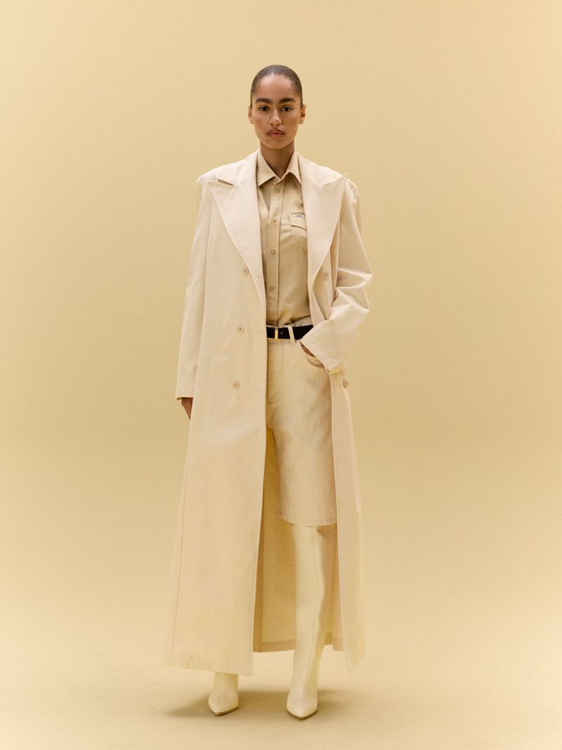 Model Erika Blanc wearing a long cream trench coat, beige shirt, matching shorts, and knee-high boots against a warm beige background.