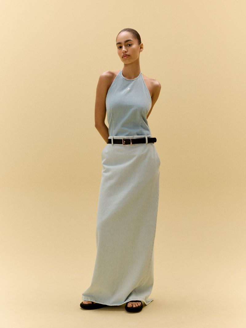 Adult woman in a halter top and long skirt stands in front of a neutral backdrop in a full shot, wearing casual sandals.