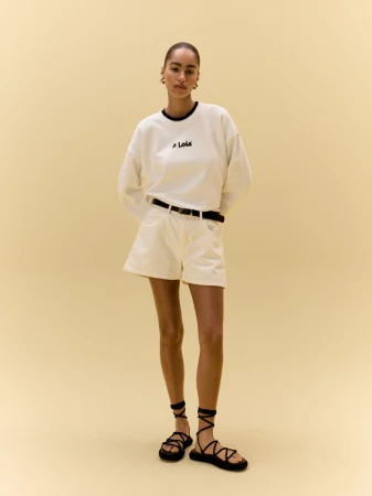 Model Erika Blanc in a white Lois logo sweatshirt, cream shorts, and black lace-up sandals standing against a warm beige backdrop.