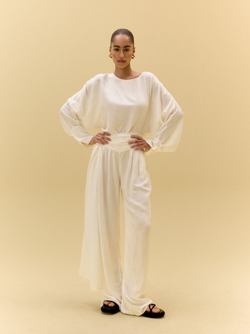 Full-length shot of a young woman in a white blouse and palazzo pants, posing in a studio with a light yellow seamless backdrop.