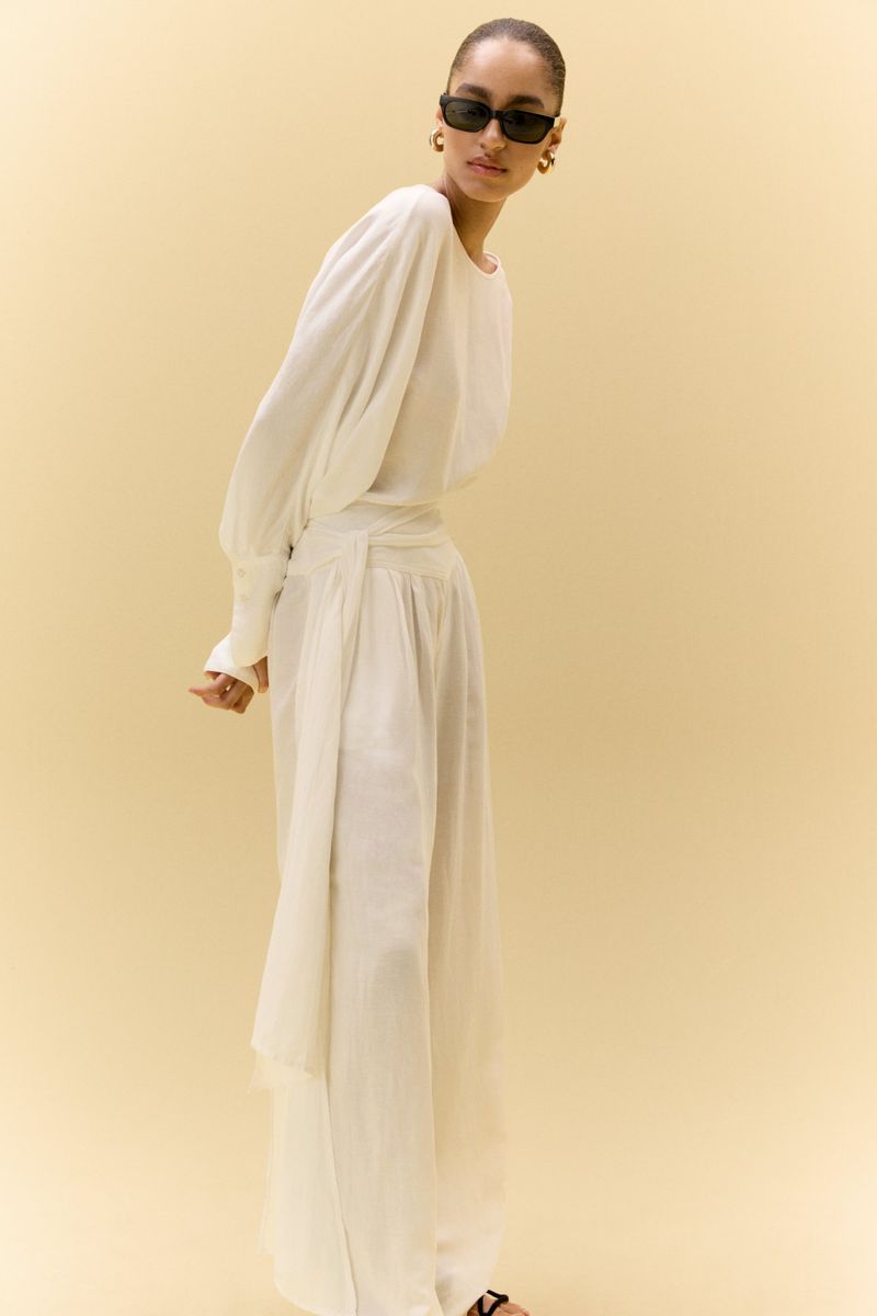 Woman in elegant white blouse and palazzo pants poses against a light background, accessorized with black sunglasses and gold earrings.