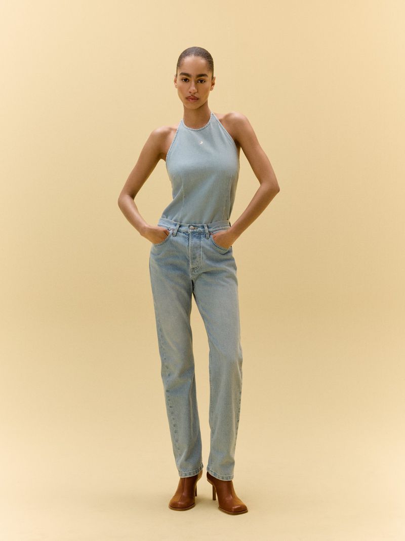 Studio shot of a woman in a denim halter top and light blue jeans, posing with hands on hips against a tan backdrop.