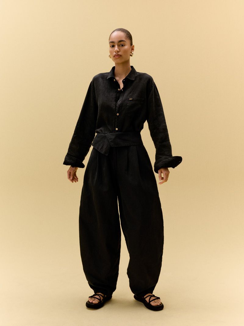Full-shot of a young adult female fashion model posing in a black button-down shirt, wide-leg pants, and flat sandals against a neutral background.