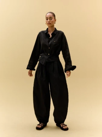 Full-shot of a young adult female fashion model posing in a black button-down shirt, wide-leg pants, and flat sandals against a neutral background.