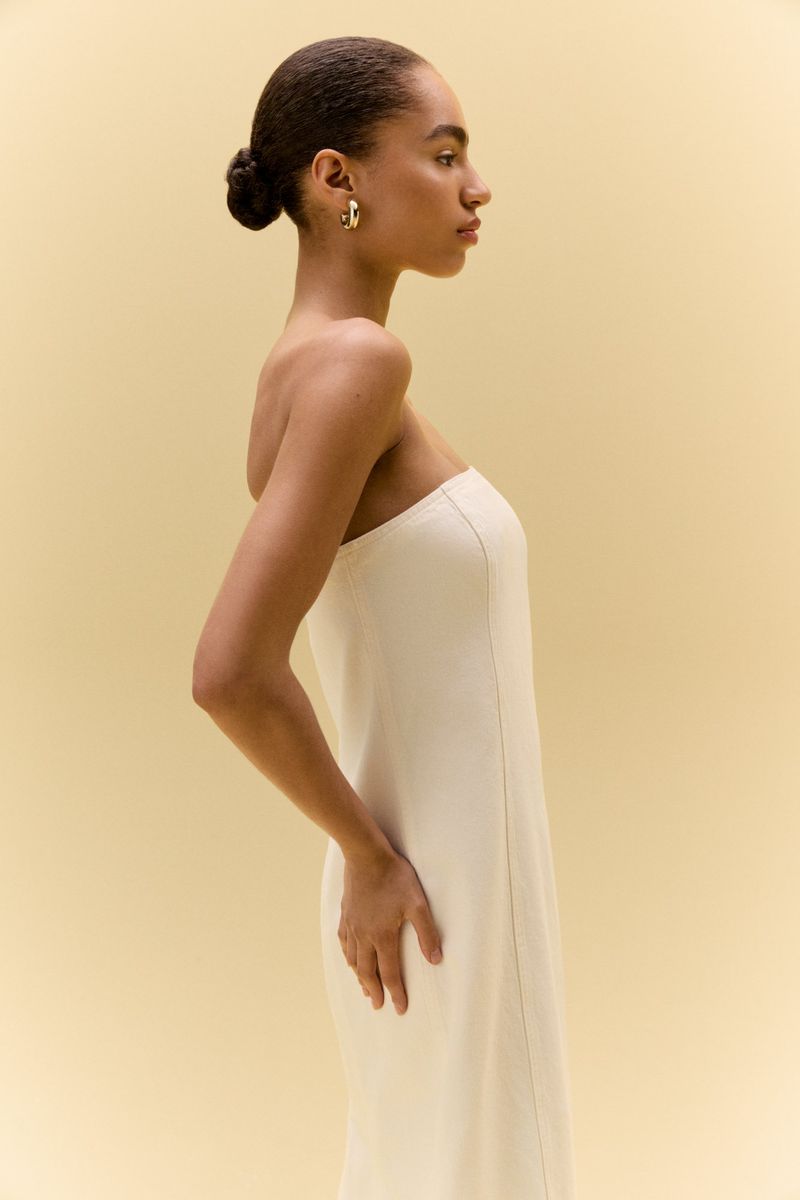 Elegant woman in profile wearing a strapless white dress and gold hoop earrings against a soft yellow background.