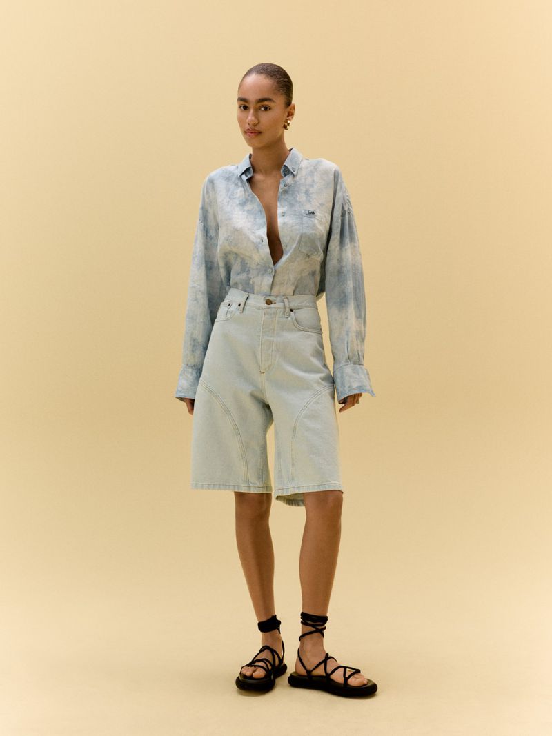 Woman in light blue tie-dye shirt and light blue denim shorts poses in front of a light yellow studio background, fashionable and chic.