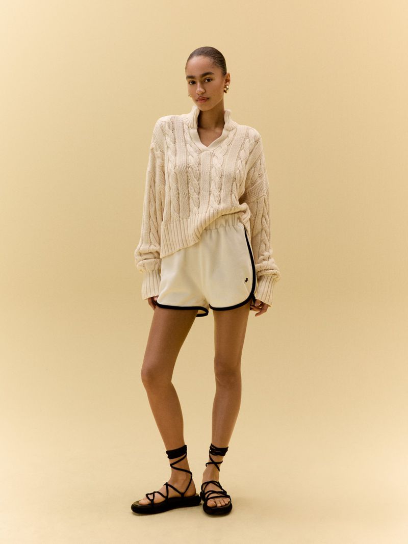Young woman poses in a relaxed, neutral indoor setting, wearing a cozy cable-knit sweater, shorts, and strappy sandals, showcasing casual fashion.