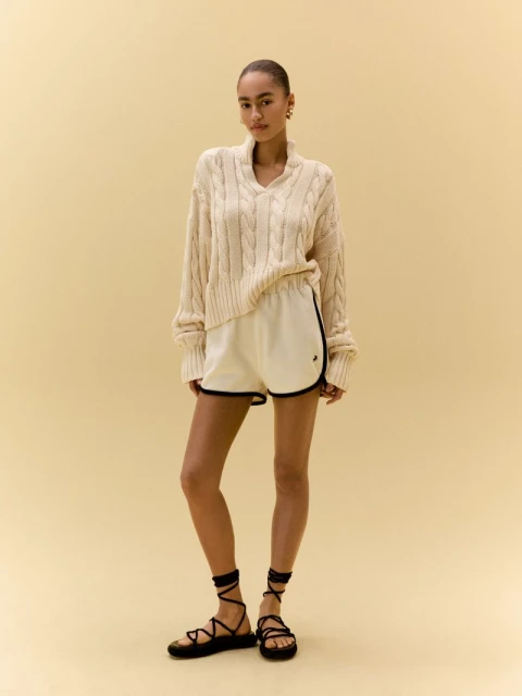 Young woman poses in a relaxed, neutral indoor setting, wearing a cozy cable-knit sweater, shorts, and strappy sandals, showcasing casual fashion.