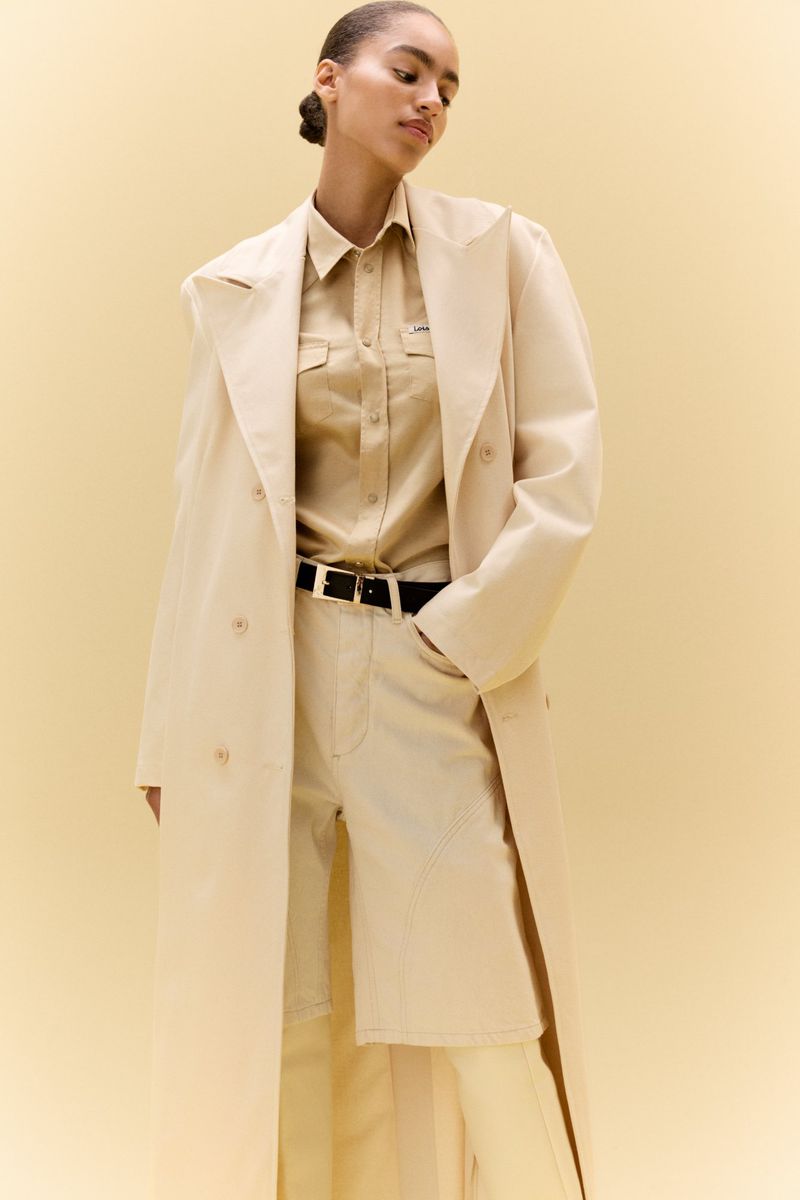 Model wearing a monochrome beige trench coat, matching button-down shirt, and tailored shorts against a cream studio backdrop.