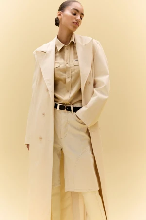 Model wearing a monochrome beige trench coat, matching button-down shirt, and tailored shorts against a cream studio backdrop.