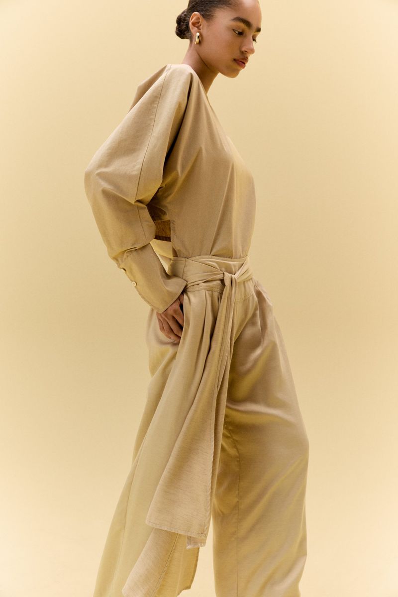 Adult female in a light-brown jumpsuit with a tie, posing in a studio with a neutral backdrop, styled hair, and natural skin.
