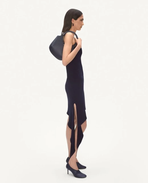 A stylish woman in a fitted black dress stands in a white studio, holding a black handbag, complemented by matching heels and dark hair.